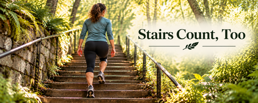 A dark-haired woman in a teal athletic top climbs a long outdoor staircase with metal railings, surrounded by sunlit trees, moss-covered stone walls, and lush greenery.