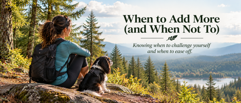 A dark-haired woman sits beside a tricolor Cavalier King Charles Spaniel on a rocky overlook, looking across a forested valley and lake. Embedded text reads “When to Add More (and When Not To)” with the subtitle “Knowing when to challenge yourself and when to ease off.”